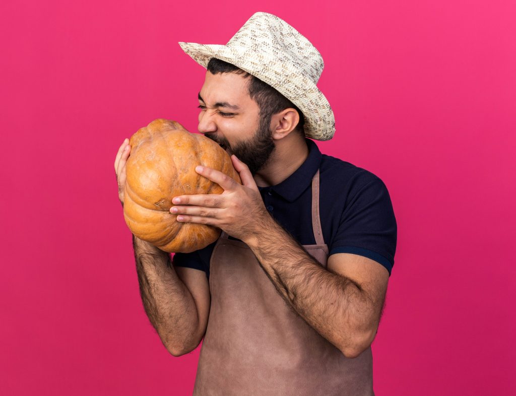 young male gardener wearing gardening hat pretending to eat pumpkin isolated on pink background with copy space