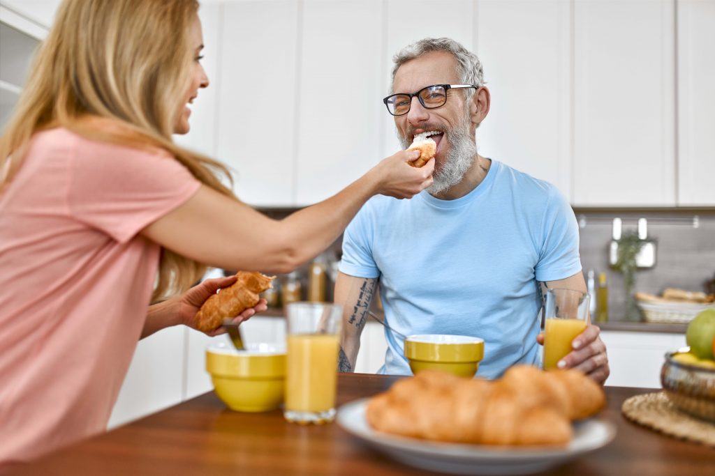 Senior couple are having breakfast. The wife feeds her husband a croissant and has fun with him early in the morning in the kitchen.