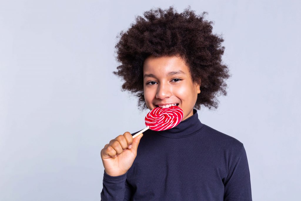 White strong teeth. Calm and smiling little man eating bright candy on wooden stick and being in great mood