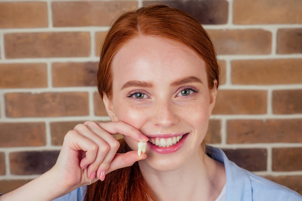 femme rousse souriante sur fond de brique tient une dent de sagesse retirée chez les dentistes de boucherville
