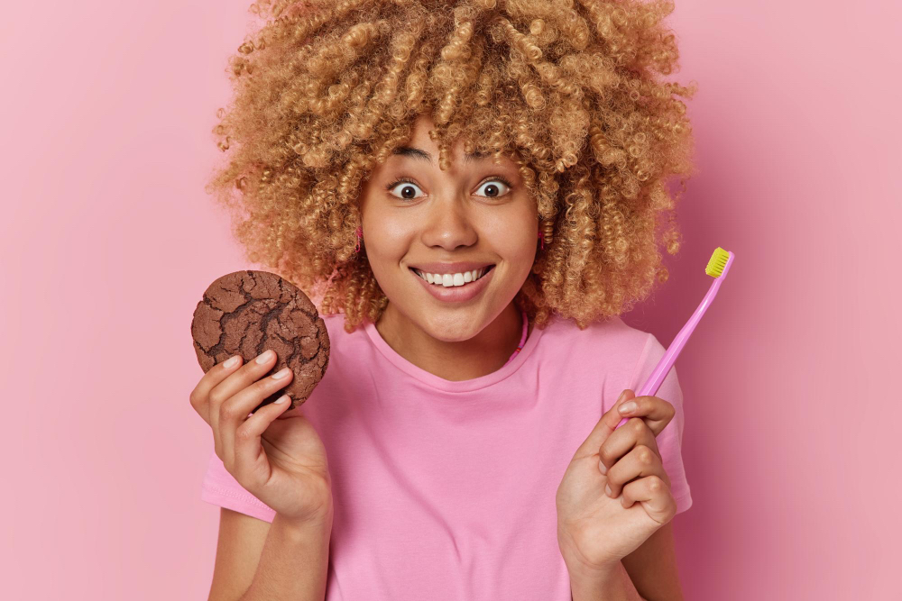 femme souriante dents blanches tenant biscuit et brosse à dente sur fond rose