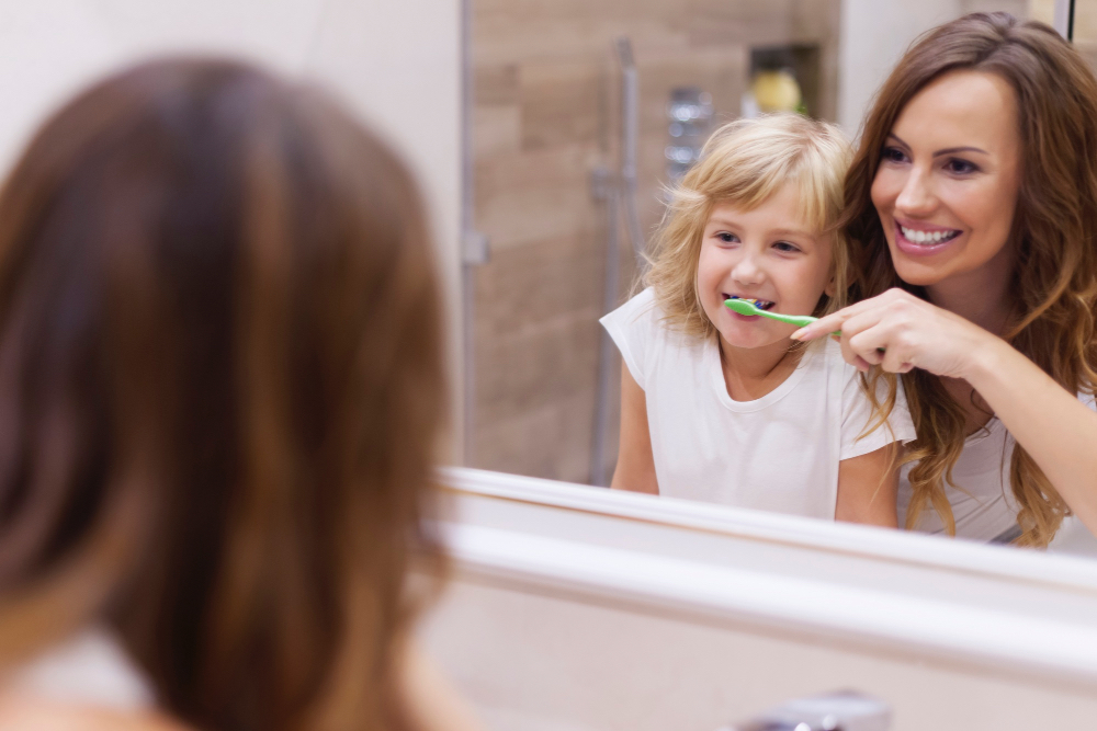 mother brushing her daughter's teeth both smiling and having fun in front of mirror